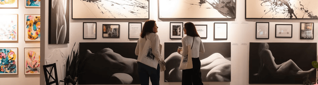 Two women viewing artwork in an art gallery, surrounded by framed paintings and photographs on the walls.